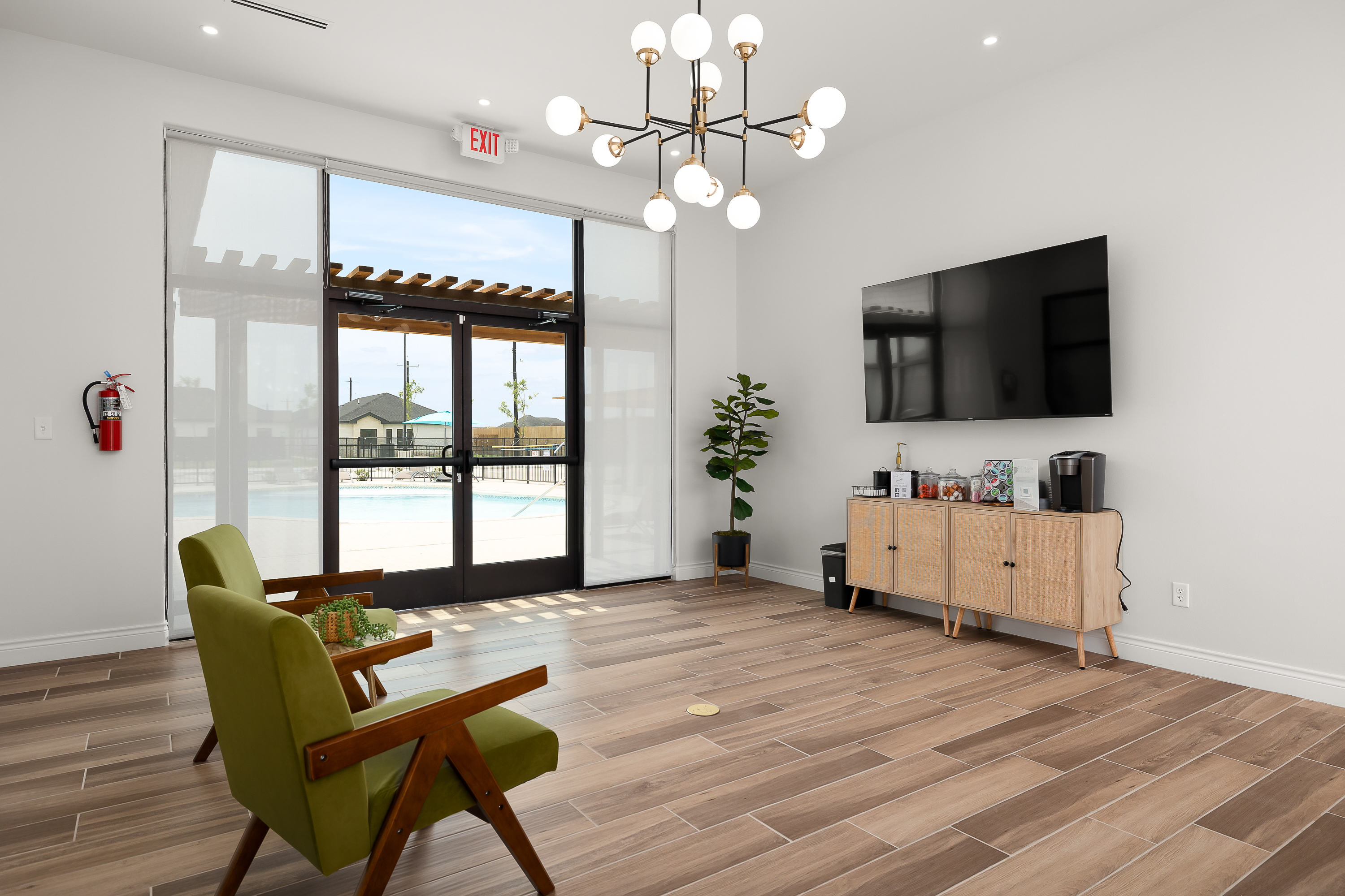 Modern living room with green lounge chairs, a wall-mounted TV, and a multi-bulb chandelier, overlooking a pool through large glass doors.