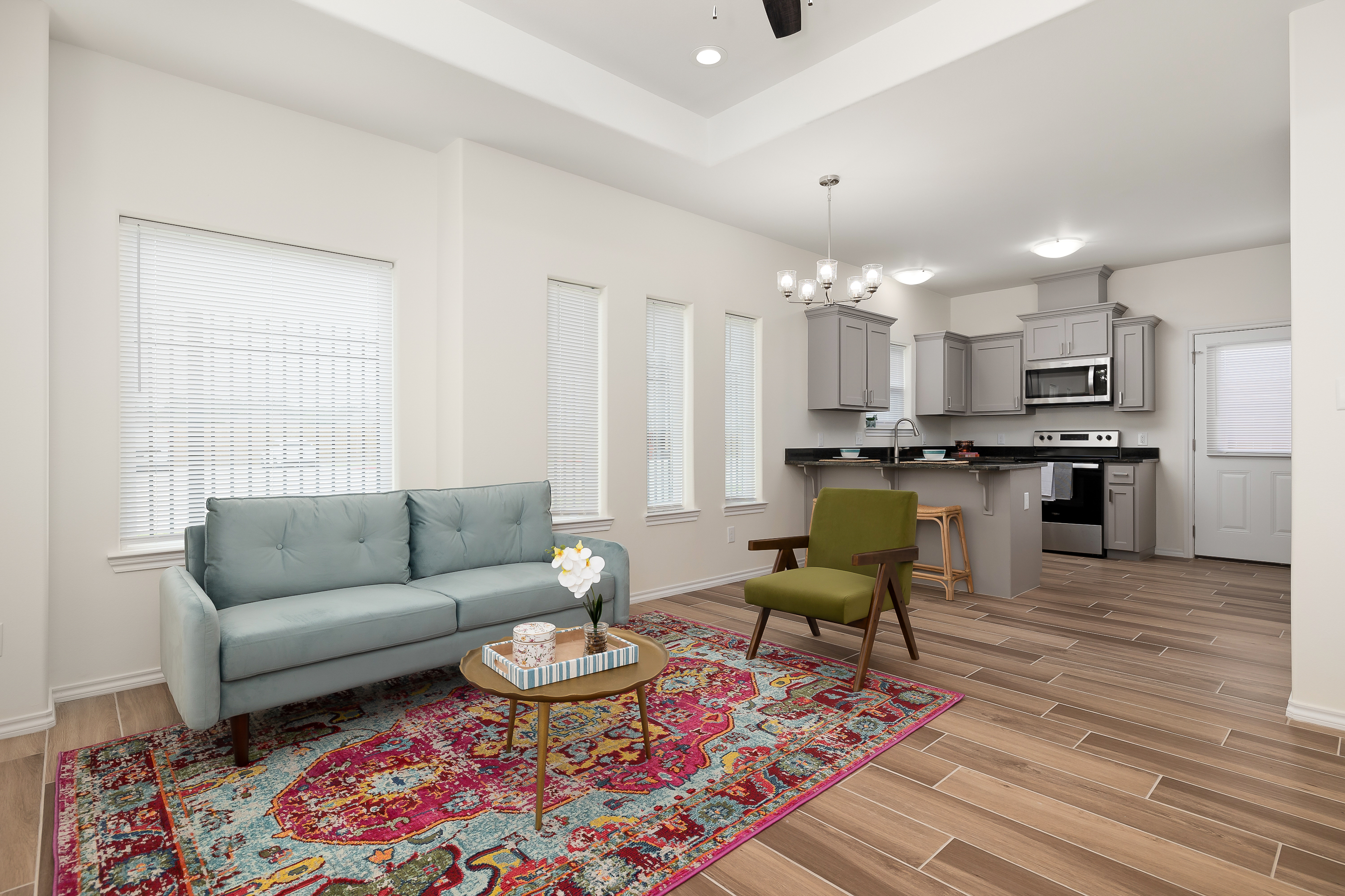 Open-concept living room with light blue sofa, colorful patterned rug, and wood-look tile flooring; view into a gray-kitchen with stainless appliances and a chandelier overhead on the island.