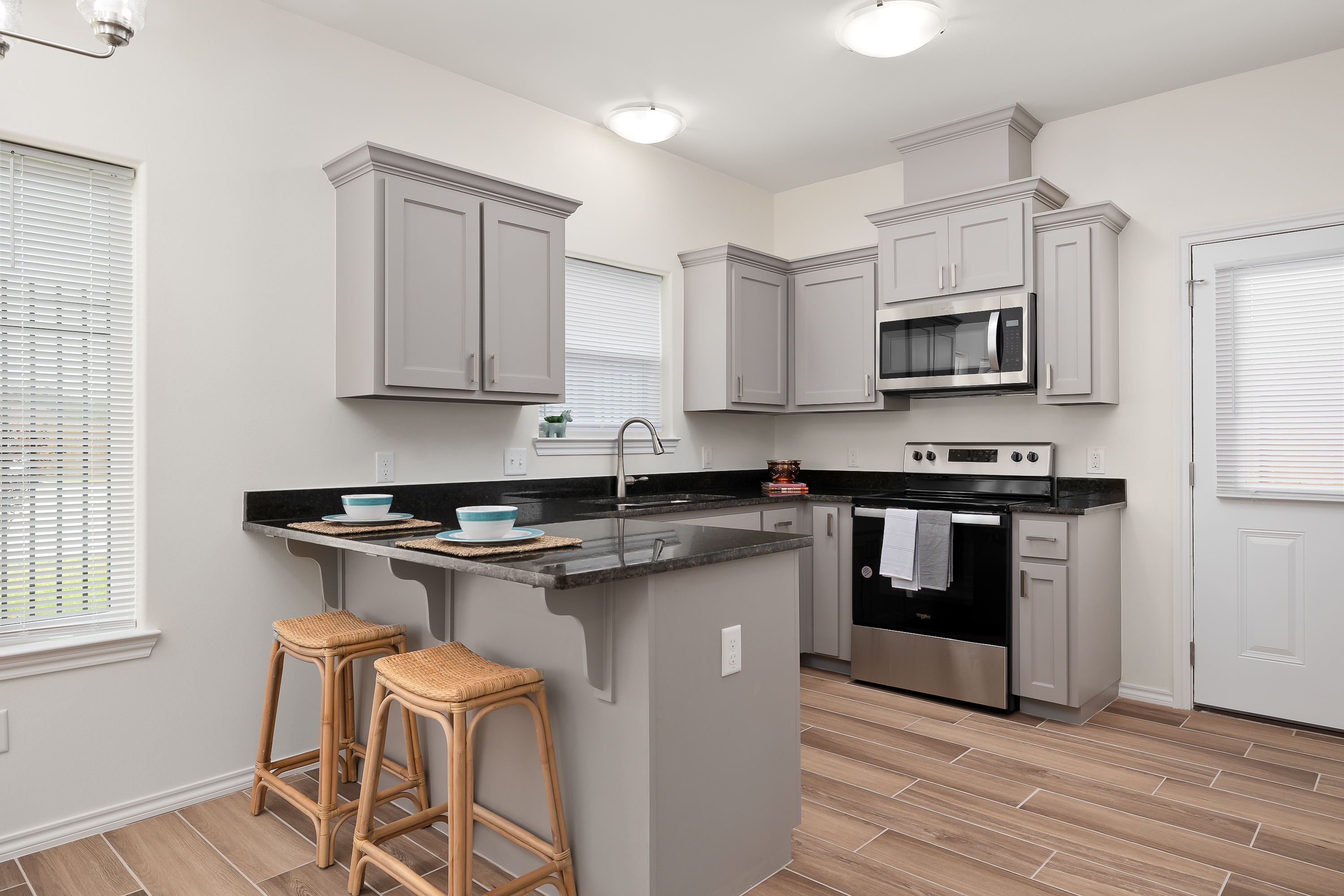 Modern kitchen with gray shaker cabinets, black countertops, and a stainless steel oven/microwave setup. A breakfast island with three wicker stools invites you to sit.