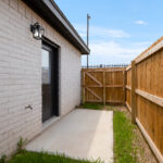 Exterior brick wall with a black door and wall-mounted lantern, alongside a wooden privacy fence and concrete walkway.