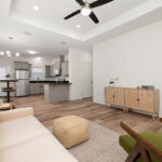 Open-concept living space with a beige sofa facing a kitchen, wooden floors, and a ceiling fan above.