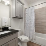 Modern gray bathroom with a granite countertop, double sink, wall cabinet, toilet, and a tiled shower with a striped curtain.