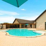 Outdoor hotel pool area with turquoise water, tan brick buildings, and teal umbrellas; lounge chairs line the pool deck, under a bright blue sky.