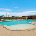 Outdoor pool area with a curved, light-blue pool, tan pool deck, and rows of lounge chairs under turquoise umbrellas.