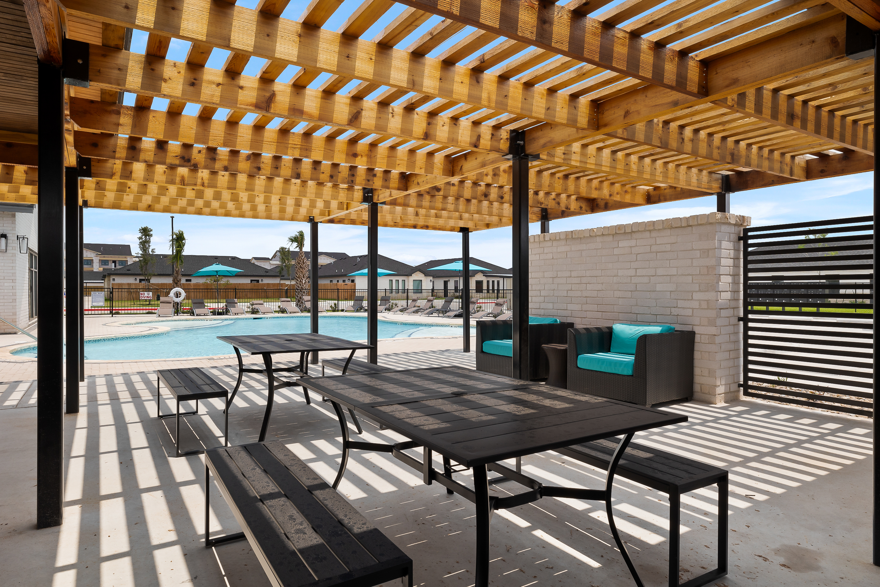 Poolside seating under a wooden pergola with teal cushions, overlooking a curved swimming pool and blue umbrellas in the background.
