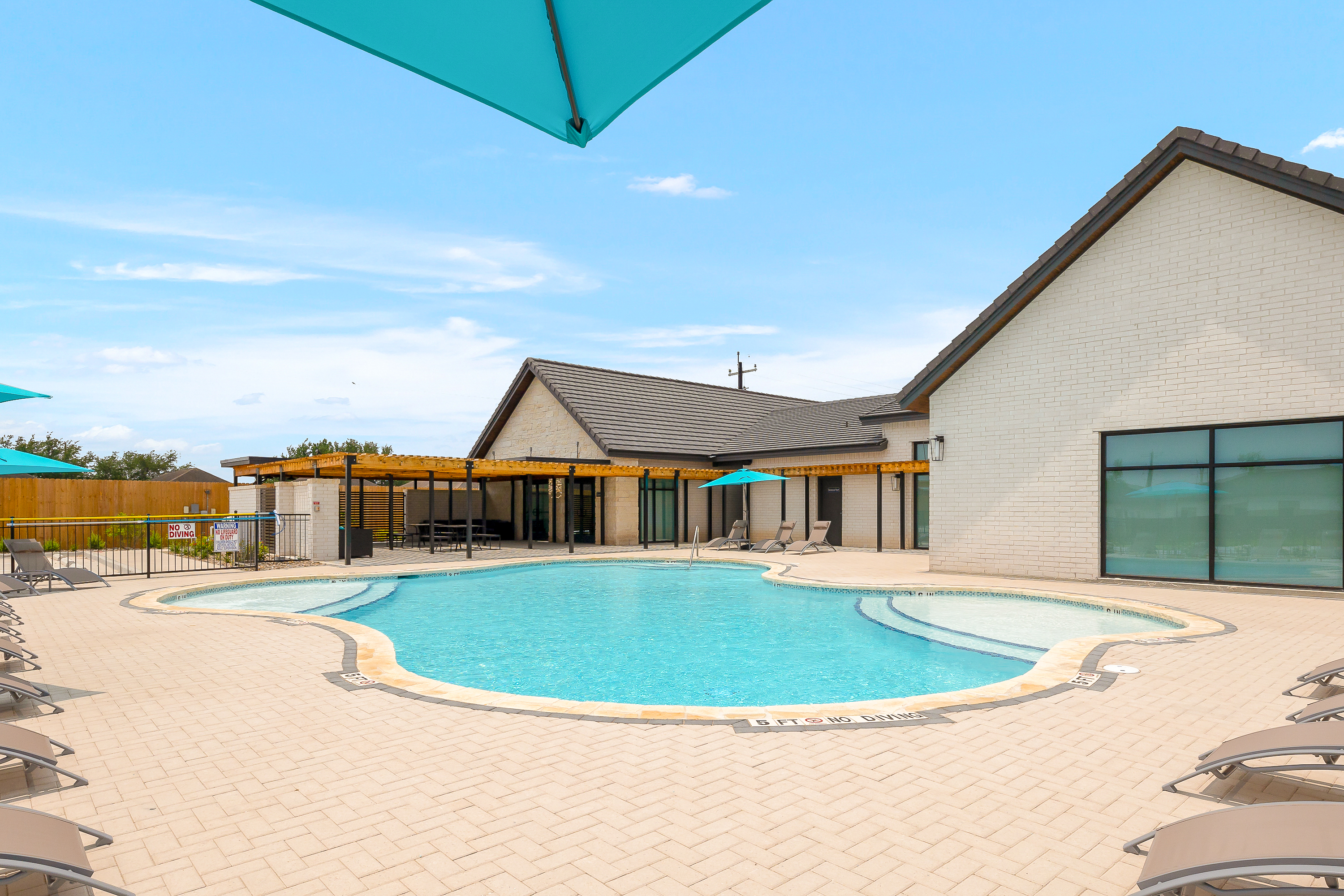 An outdoor pool area with lounge chairs, turquoise shade sails, and a light brick building in the background under a clear blue sky, no diving signs visible.
