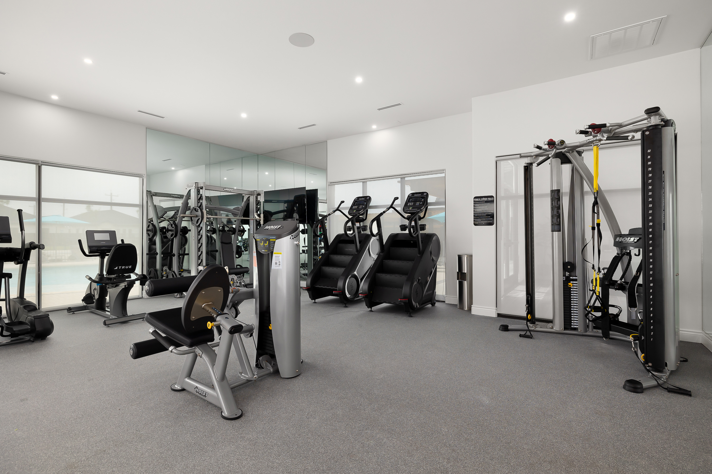 Bright modern gym with row of cardio machines (treadmills and ellipticals) in the foreground and weight machines along mirrored wall in background. Large windows provide natural light.