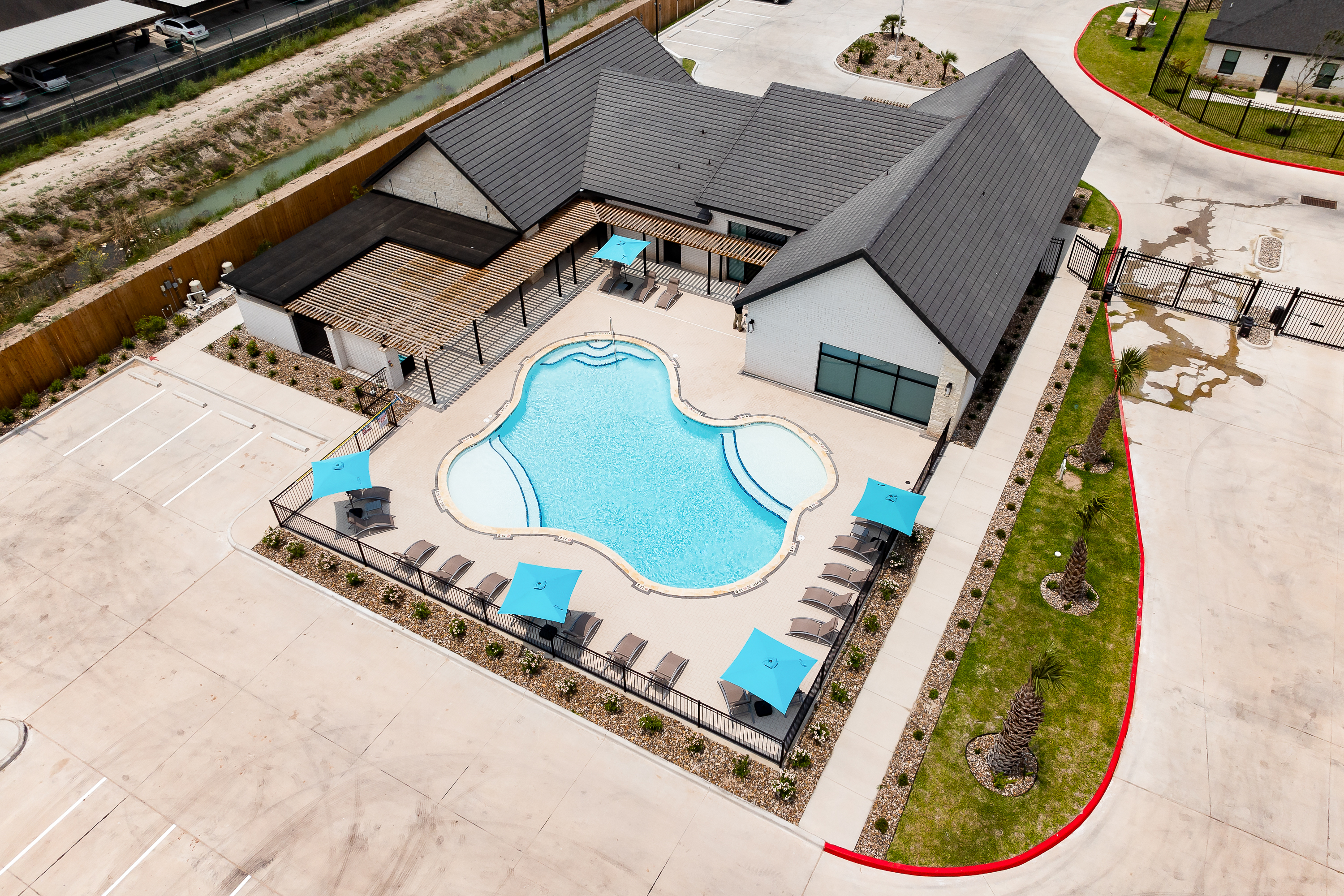 Aerial view of a resort-style pool area with a curved blue pool, lounge chairs, and blue umbrellas beside a white brick building with a dark roof.
