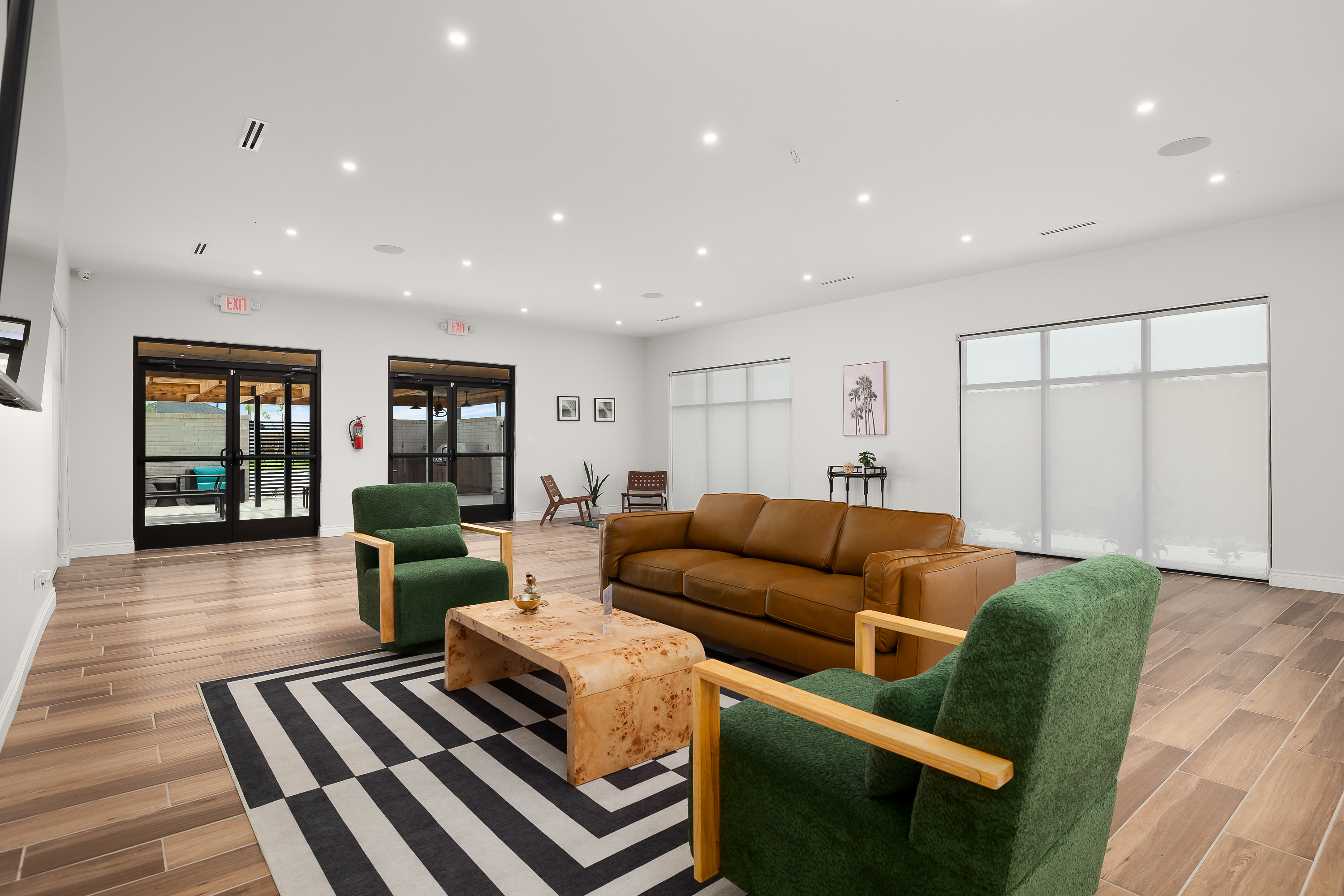 Bright modern lounge with green upholstered chairs, a tan leather sofa, and a wooden coffee table on a black-and-white striped rug; large windows and glass doors lead outside.