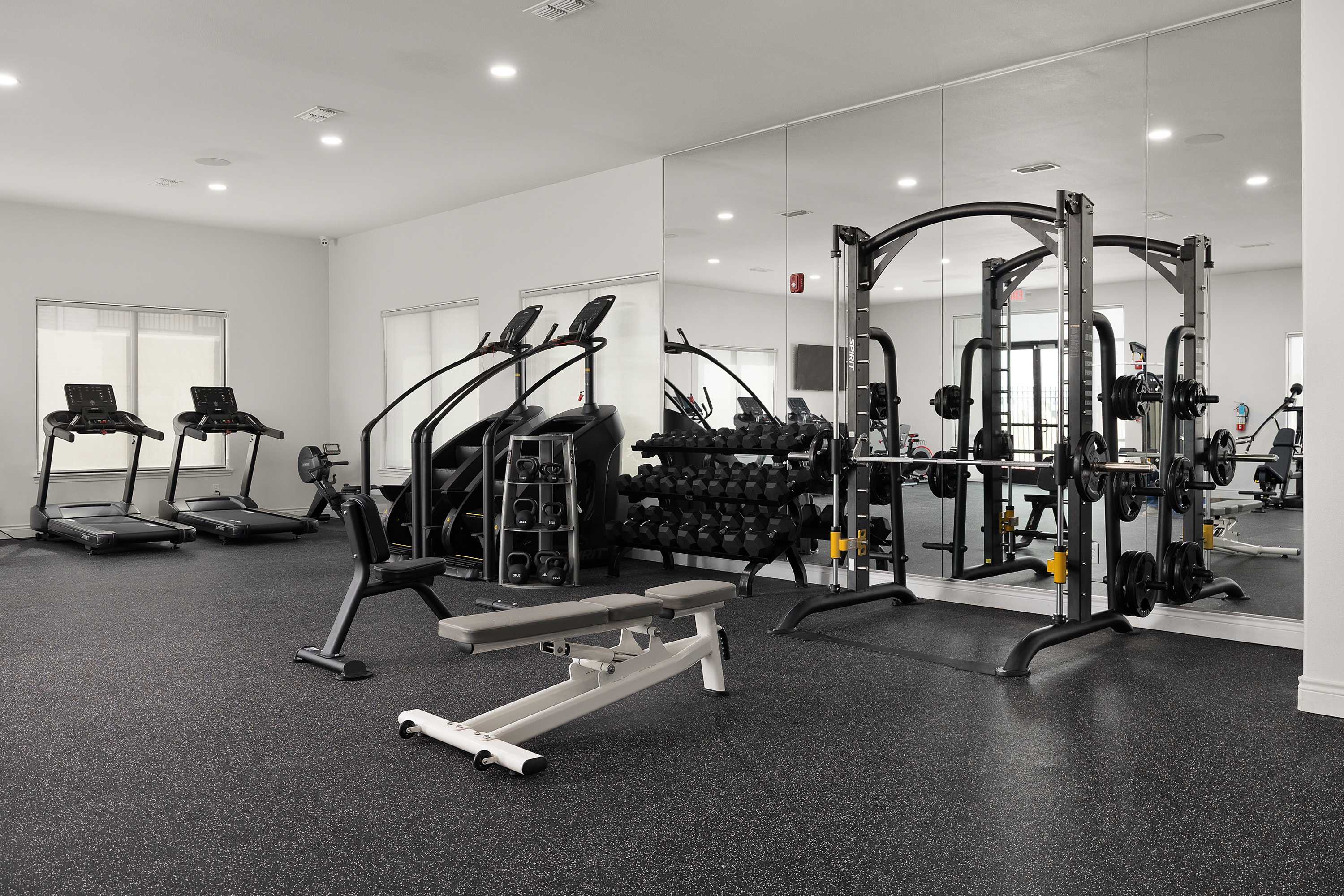 Bright gym interior with two treadmills on the left and a large free-weight rack and squat station on the right, mirrored wall in back.