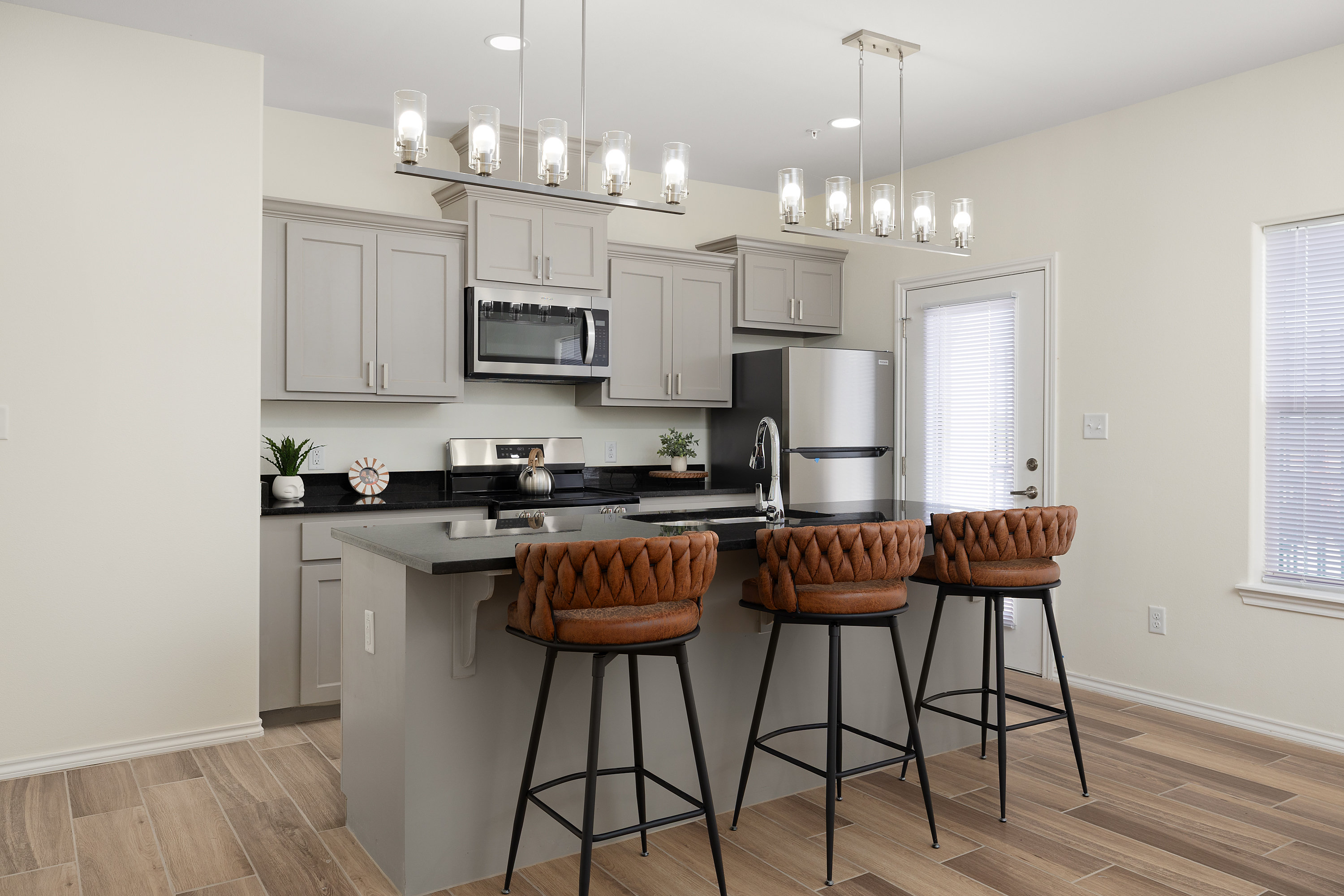 Modern kitchen with gray cabinets, stainless steel appliances, and a stone island surrounded by four tufted brown bar stools under two pendant-light fixtures.