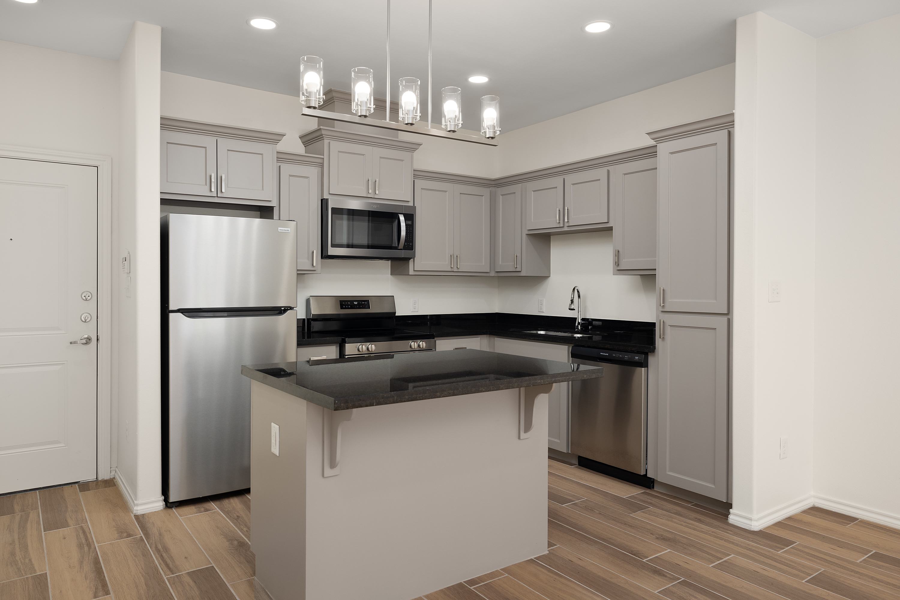 Modern kitchen with gray cabinets, stainless steel fridge, microwave, and a dark countertop island under a contemporary pendant light fixture on a white wall.
