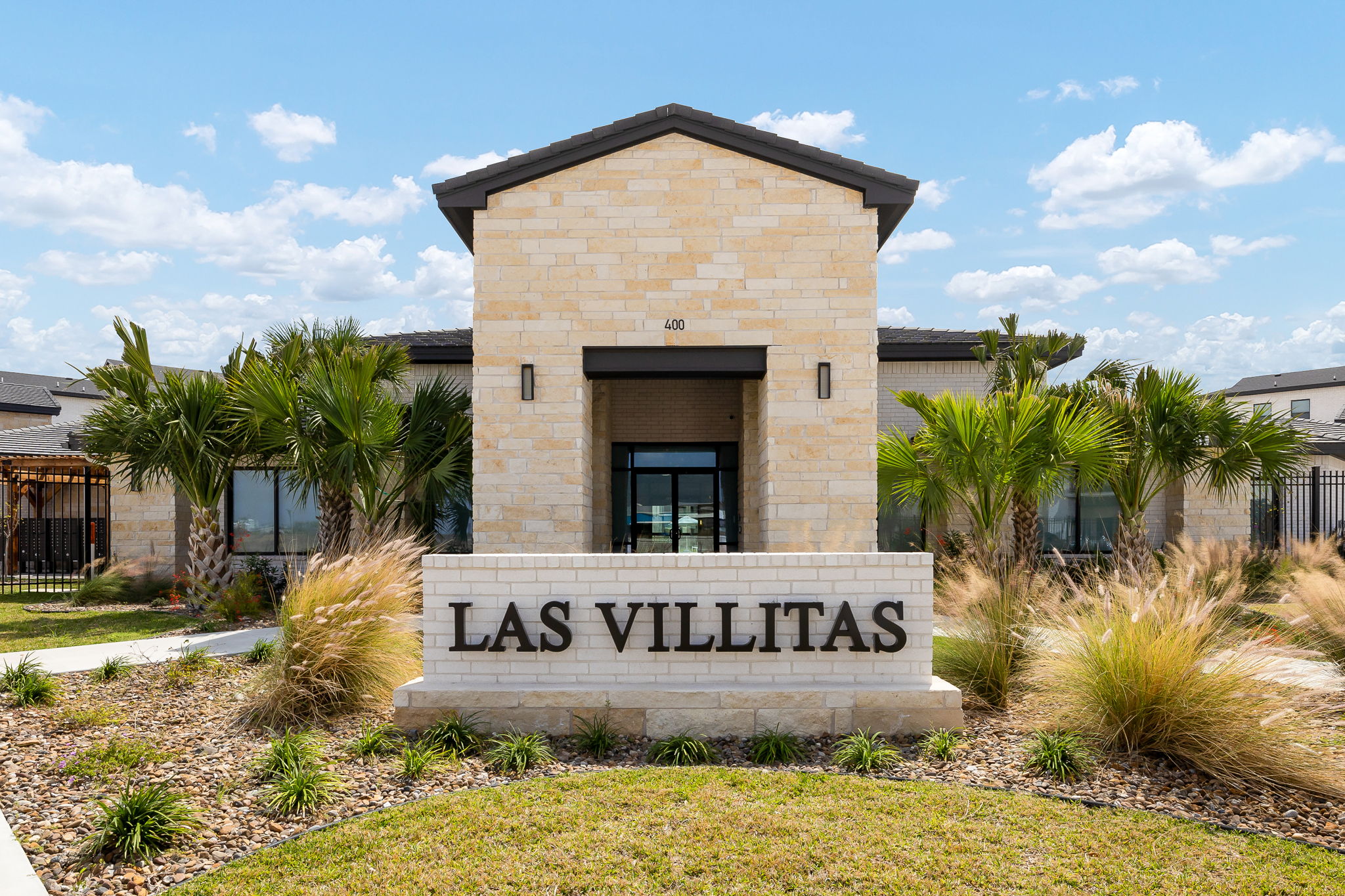 Stone entrance sign reading LAS VILLITAS with a beige brick tower, palms, and clear blue sky behind.