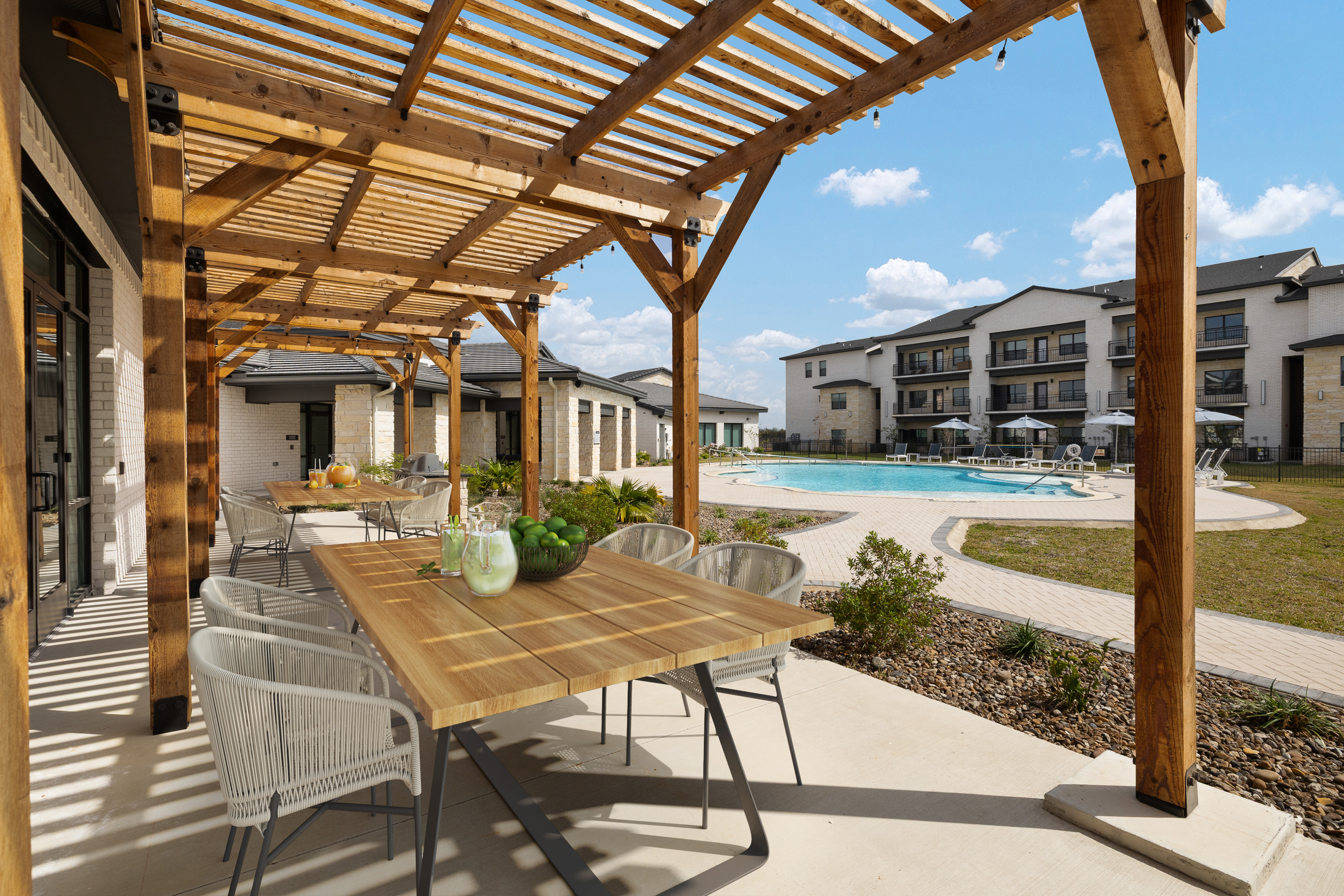 Outdoor dining area under a wooden pergola beside a pool, with tables set and apartment buildings in the background on a sunny day.