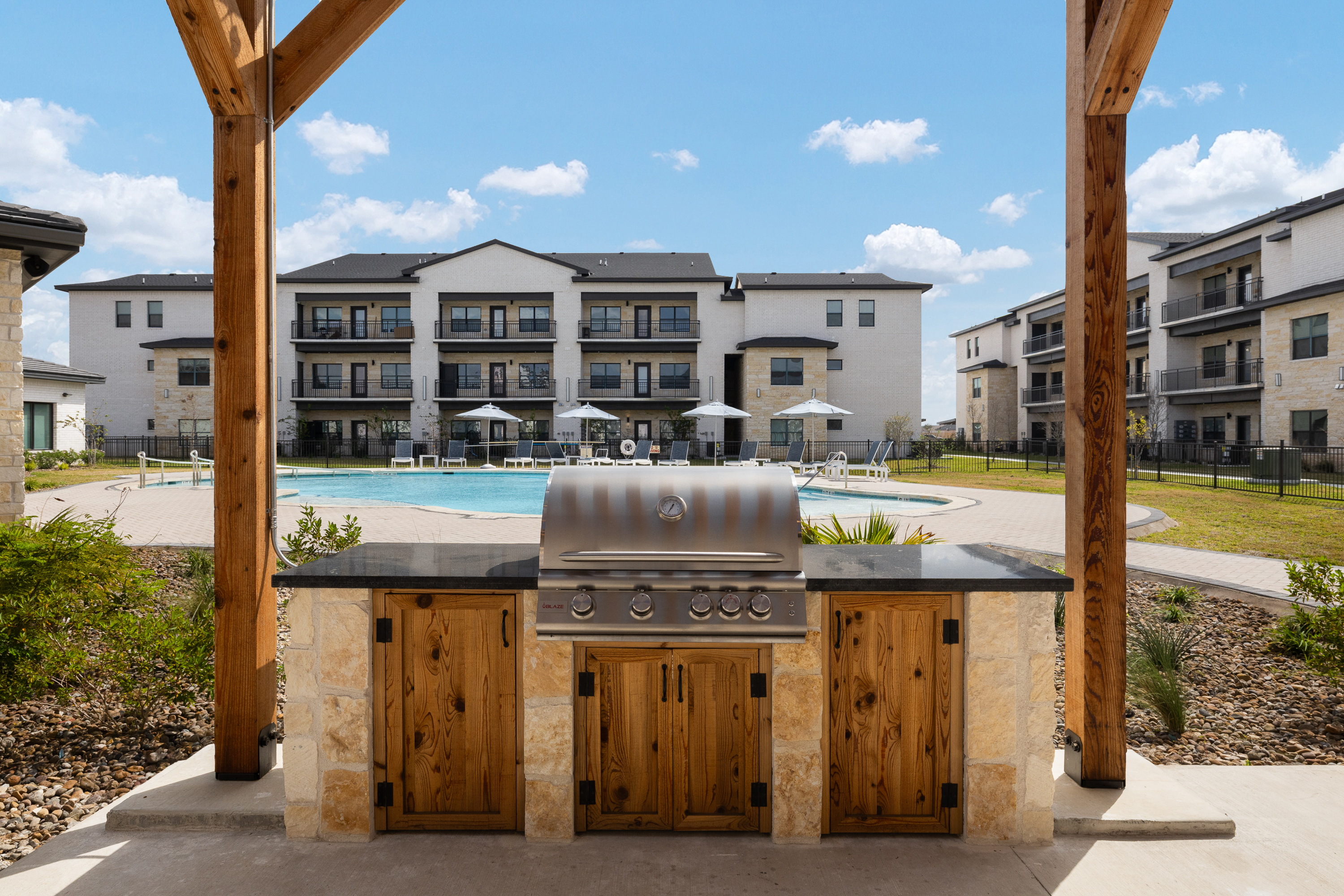 Outdoor grilling station with a stainless steel barbecue under a wooden pergola, pool and modern apartment buildings in the background.
