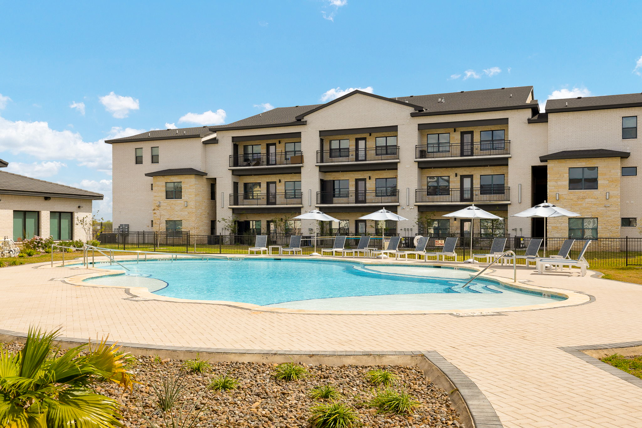 Modern apartment complex with a curved blue pool in the foreground, surrounded by lounge chairs and umbrellas under a clear sky.