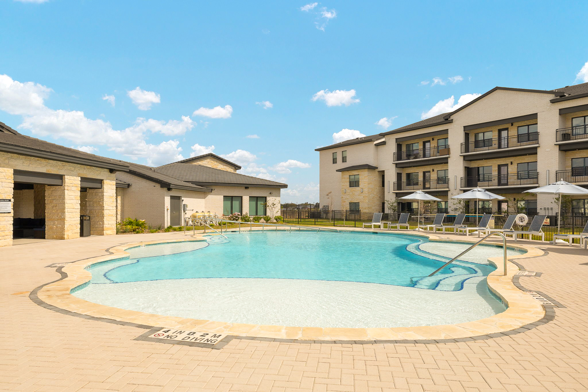 Outdoor pool area of a residential complex with beige stone-patio, lounge chairs, umbrellas, and multi-story beige brick buildings under a blue sky .