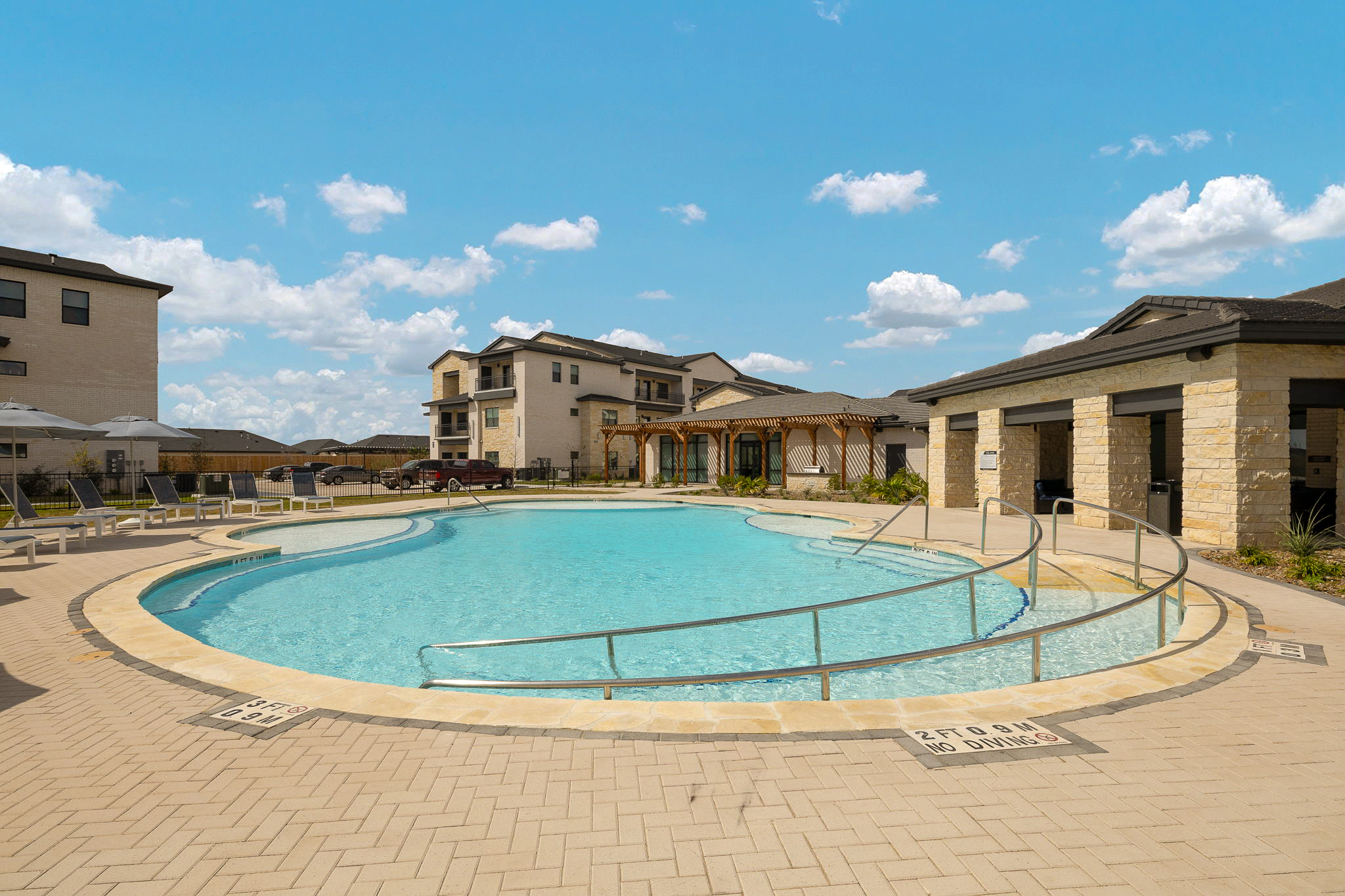Outdoor pool area of a residential complex with lounge chairs, umbrellas, and beige stone buildings under a clear blue sky.