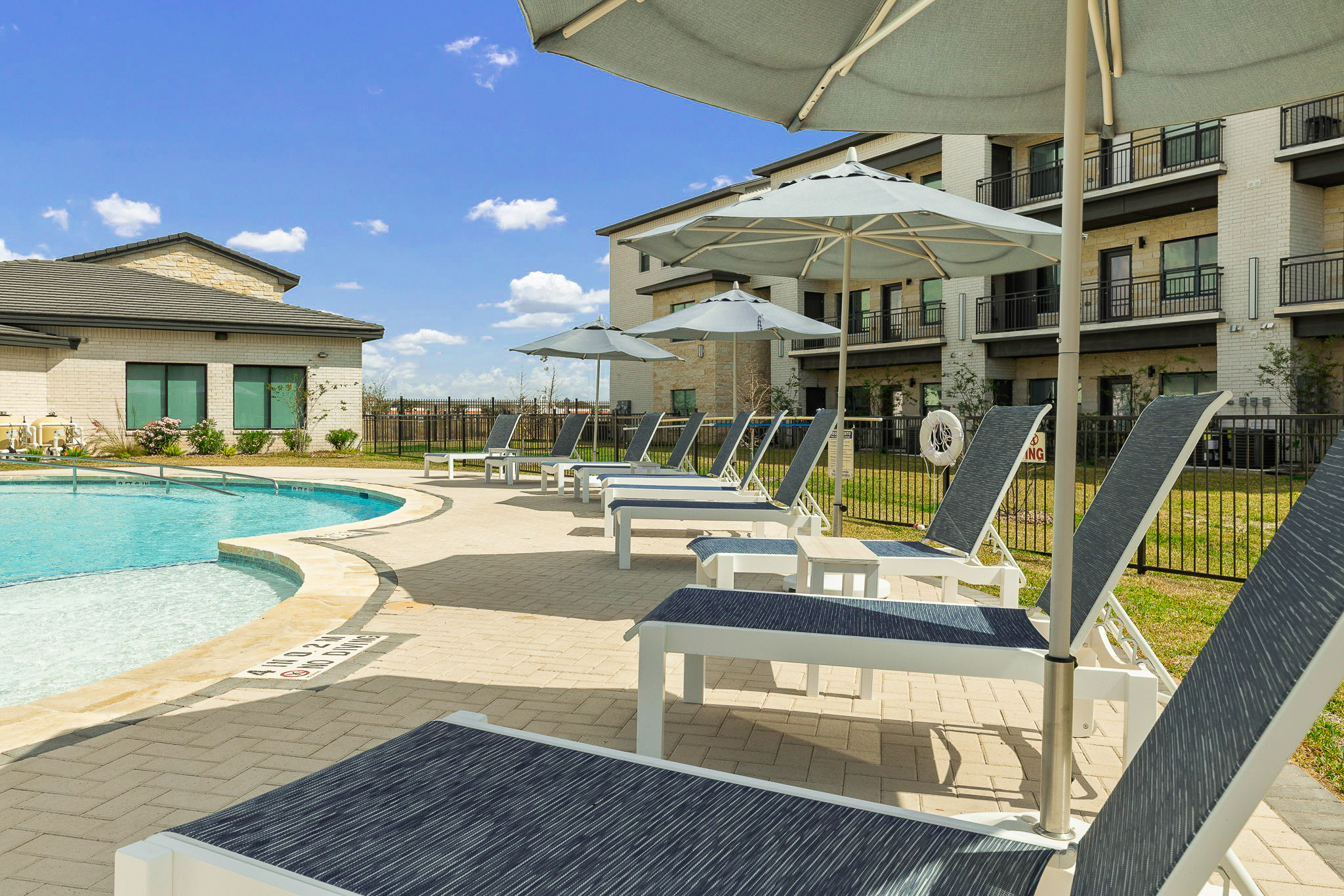 Poolside at a residential complex: loungers lined up under large umbrellas beside a curved pool, with a multi-story building in the background.