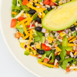 Colorful mixed salad in a white bowl with lettuce, avocado half, corn, cheese, beans, seeds, and diced vegetables.