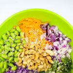 Green bowl holding a colorful veggie salad: edamame, shredded carrot, red onion, purple cabbage, sesame seeds, peanuts, and herbs on a bright background that emphasizes freshness.