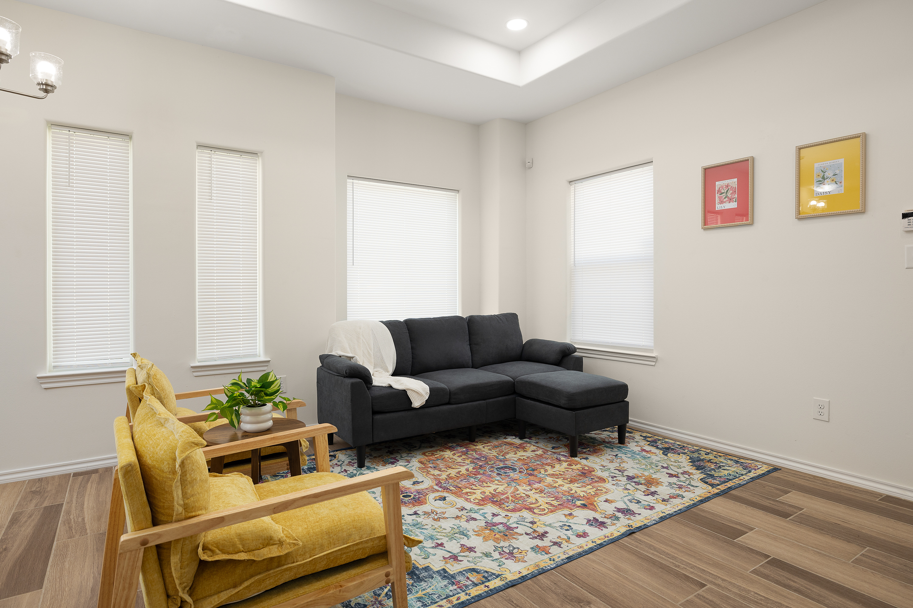 Bright living room with white blinds, a dark gray sectional sofa, and mustard chairs around a small wooden table with a plant; colorful rug adds pattern.