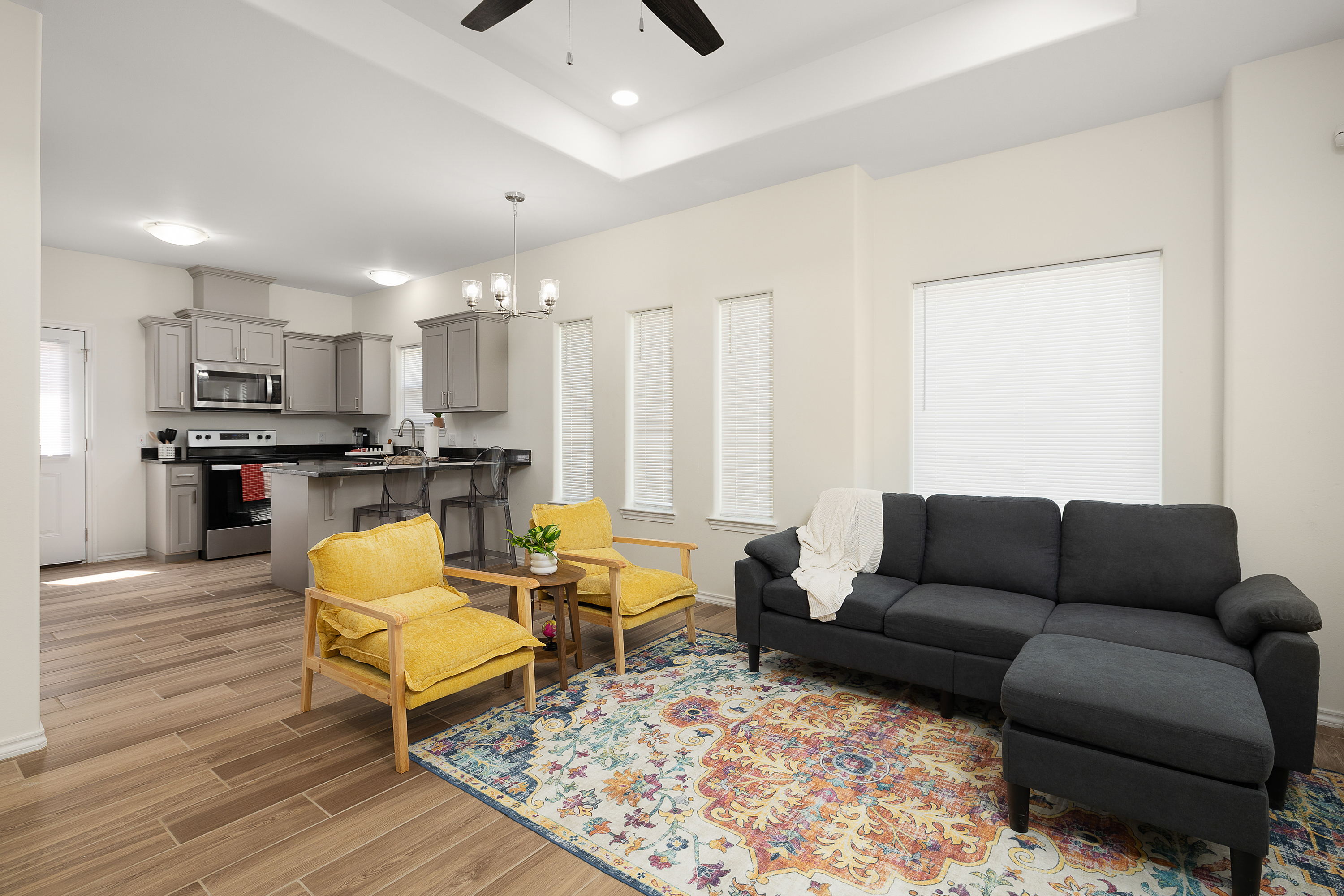 Open-concept living room and kitchen with gray cabinets, stainless-steel appliances, a kitchen island with yellow chairs, and a colorful rug.