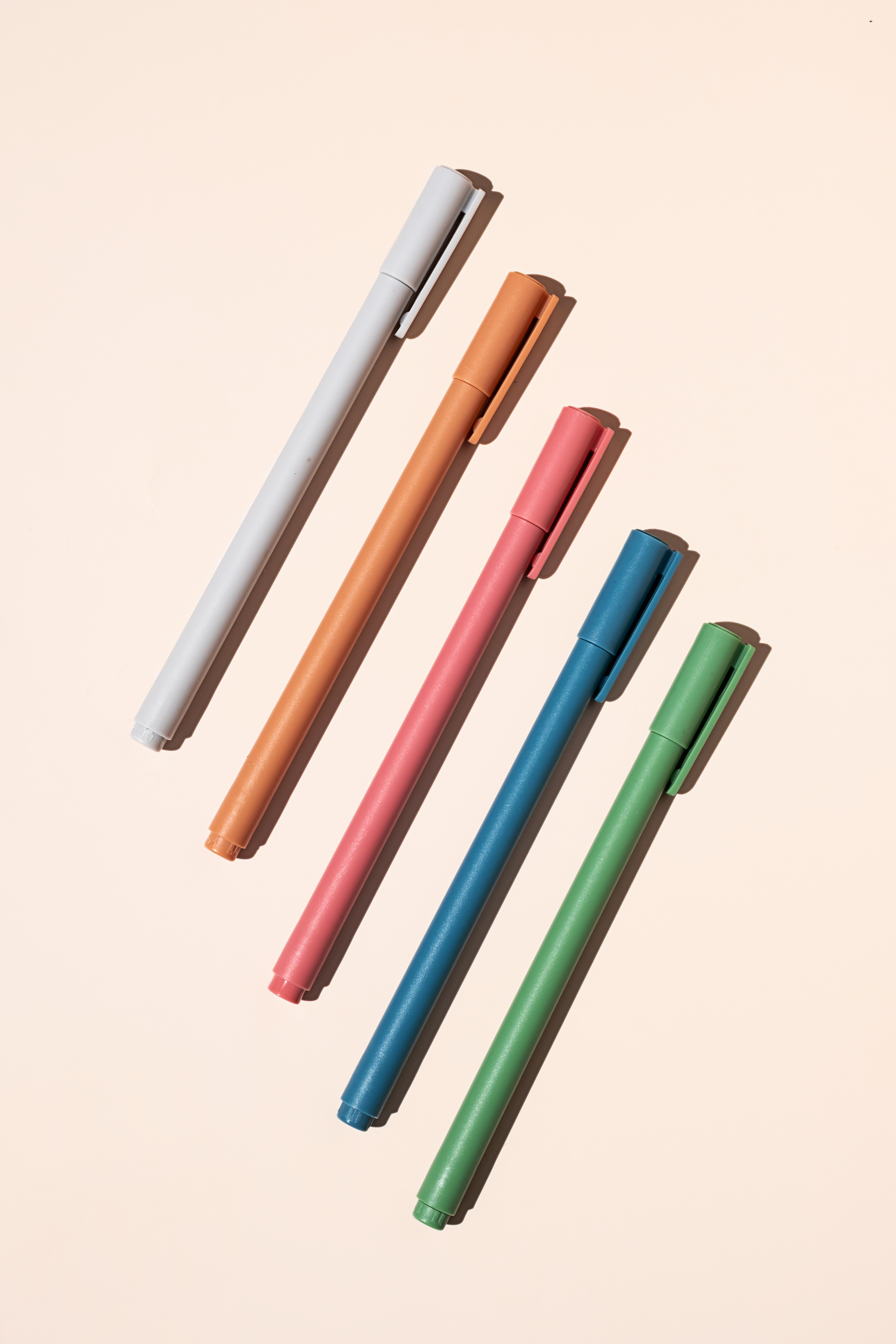 Six pastel-colored marker pens arranged diagonally on a peach background with shadows behind them.
