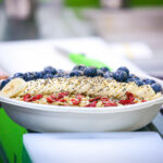 Gloved hand presenting a dessert bowl topped with blueberries, sesame seeds, granola, and crushed berries.
