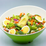 Colorful salad in a green bowl featuring hard-boiled egg slices, avocado, broccoli, shredded carrots, lettuce, and red onion.