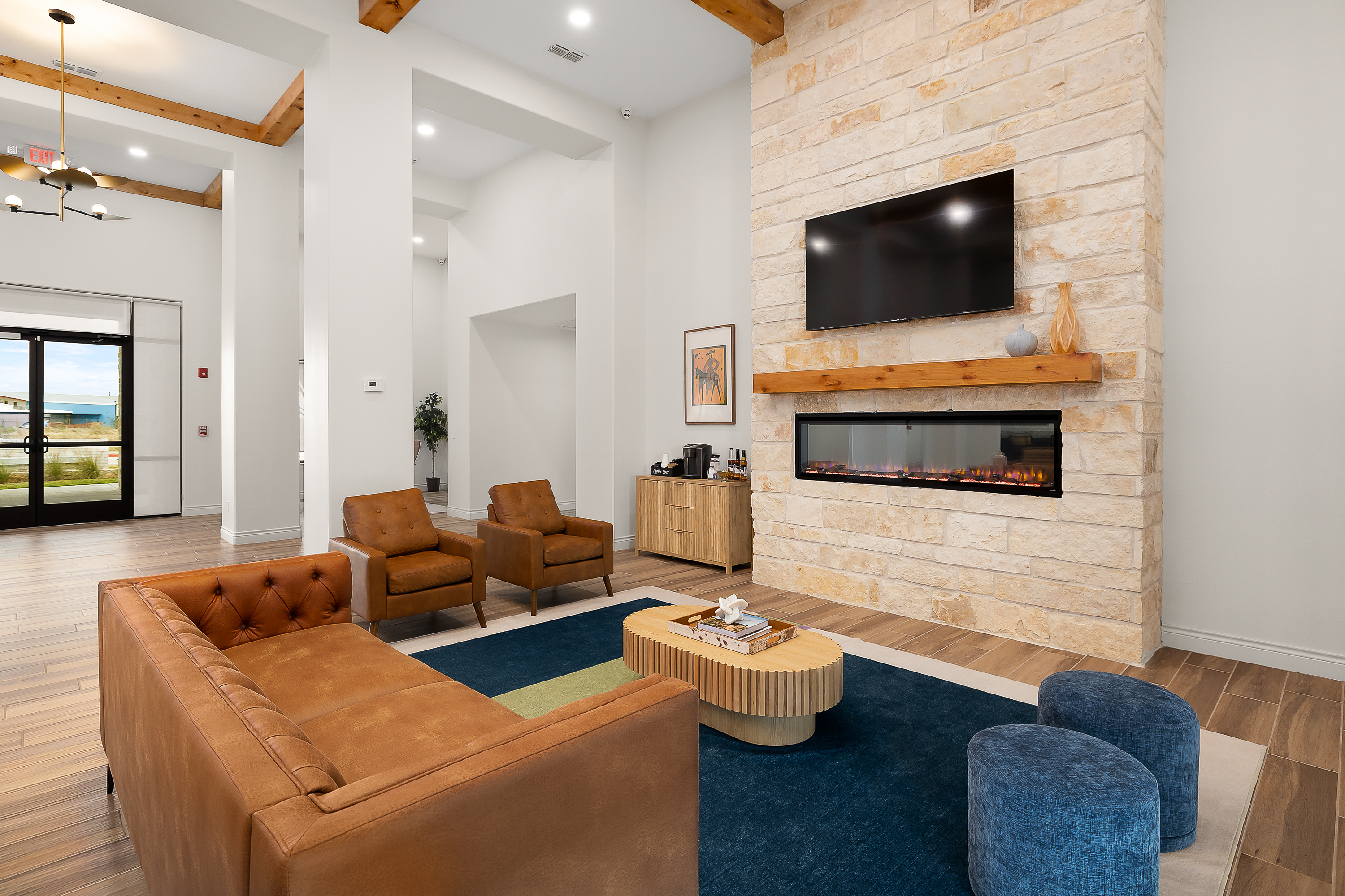 Modern living room with brown leather sofas around a round wooden coffee table, dark blue and multicolor rug, and a stone fireplace with a mounted TV.