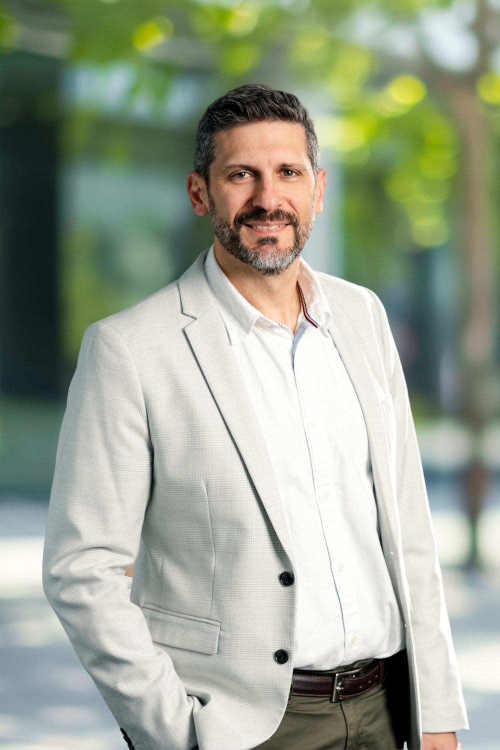 Smiling man in a light gray blazer and white shirt posing outdoors with green, sunlit foliage in the background.