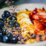 Close-up of a fruit yogurt bowl with blueberries on the left, banana slices in the center, and sliced strawberries on the right, topped with granola and orange zest/crumbs.