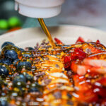 Close-up of a dessert bowl with blueberries, strawberries, crushed nuts, and caramel being poured over the fruit.