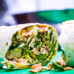 Half-open wrap with shredded chicken, lettuce, and herbs spilling from a soft tortilla onto a green surface.