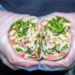 Two halves of a wrap filled with greens, chicken, tomatoes, onions, and cheese, held by gloved hands in pink gloves.