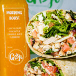 Bottle of Morning Boost orange juice with green cap beside a veggie chicken wrap filled with greens and diced tomatoes on a white surface, Goji branding in background.