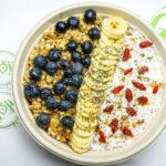 Overhead view of a breakfast bowl topped with blueberries, sliced banana, granola, shredded coconut, goji berries, and chia seeds without text in frame.
