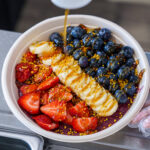 Colorful fruit bowl with blueberries, strawberries, sliced bananas, and bee pollen on a purple smoothie base with drizzle on top.