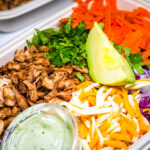 Burrito bowl toppings: shredded beef, avocado wedge, cilantro, shredded carrots, chopped onions, and green sauce in a takeout tray.