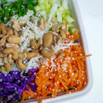 Colorful salad bowl with shredded carrots, purple cabbage, greens, sesame seeds, and chopped nuts in a white container.