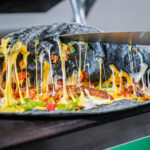 Close-up of a cheese-stretching pizza with peppers and vegetables being lifted by a metal spatula on a hot griddle.