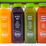 Row of four juice bottles with green caps in orange, purple, olive green, and red labels, reading Morning Boost, UN-BEETABLE, GREEN-ERGYLE, and GINGER LEMON CLEANSE.