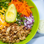 Colorful salad bowl with avocado half, shredded cheese, carrots, peppers, onions, cilantro, and crispy chicken, plus a dipping sauce on the side.