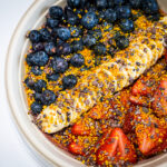 Bowl of yogurt topped with blueberries, banana slices, strawberry pieces, bee pollen, and chocolate shavings.