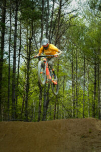 Cyclist in an orange shirt wearing a helmet mid-air on a dirt jump in a forest trail