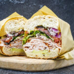 Halved deli sandwich with shredded turkey, greens, red onion, and tomato on a crusty bun, wrapped in parchment on a wooden board.