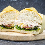 Two halves of a sandwich with a creamy filling and greens on a round wooden board against a dark background, shown in cross-section.