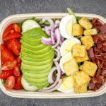 Colorful salad bowl with avocado slices, tomatoes, cucumber, red onion, hard-boiled eggs, croutons, and diced meat on greens.