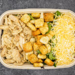 Takeout container with creamy chicken Alfredo on the left, crouton-topped greens in the center, and shredded cheese on the right.